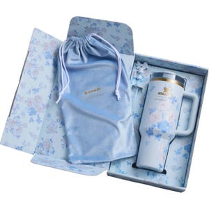 May include: A light blue Stanley tumbler with a floral pattern, a matching drawstring pouch, and a straw, all presented in a floral-patterned gift box. The tumbler features the Stanley logo and the text LoveShackFancy printed on the front surface.