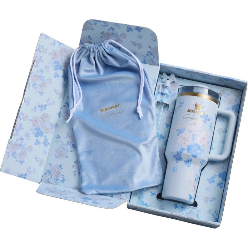 May include: A light blue Stanley tumbler with a floral pattern, a matching drawstring pouch, and a straw, all presented in a floral-patterned gift box. The tumbler features the Stanley logo and the text LoveShackFancy printed on the front surface.