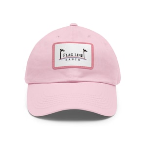 Puede incluir: Una gorra de béisbol de color rosa claro con un parche rectangular blanco en la parte delantera. El parche muestra el texto FLAG LINE RANCH en letras negras, junto a dos iconos de banderas negras y una fina línea curva roja y azul.