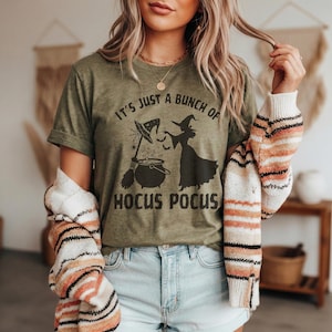 Peut inclure: Un t-shirt à manches courtes vert olive avec une impression graphique noire représentant une sorcière, un balai et un chaudron, accompagné du texte "IT'S JUST A BUNCH OF HOCUS POCUS" en caractères gras. Porté avec un gilet rayé à manches longues.