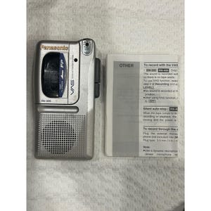 May include: A silver Panasonic RN-305 microcassette recorder with a built-in speaker, a tape compartment, and a microphone port. Beside it lies a white instruction manual with black text detailing recording functions and silent auto-stop.