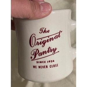 May include: A white ceramic mug featuring dark red text that reads The Original Pantry, SINCE 1924, and WE NEVER CLOSE. The mug has a smooth, glossy finish and a sturdy handle on the right side, displaying a classic diner-style design.