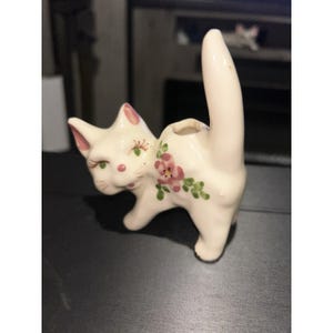 May include: A cream-colored ceramic cat figurine with a tall, upright tail and a small circular opening on its back. The cat features hand-painted pink inner ears, green eyes with long eyelashes, a pink nose, and a floral pattern on its side.