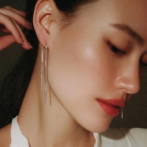 May include: A pair of silver-toned threader earrings featuring a delicate chain design that loops through the earlobe. Each earring consists of a thin, polished metal chain ending in a slender, straight bar component that hangs vertically.