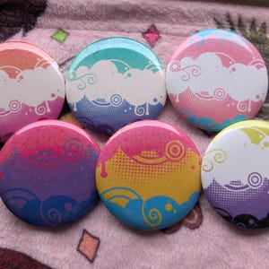Op de afbeelding: Zes ronde buttons met een herhalend grafisch ontwerp van witte gestileerde wolken, wervelende lijnen en concentrische cirkels. Elke button heeft een unieke kleurverloopachtergrond, waaronder oranje-roze, groenblauw-paars, roze-blauw, paars-blauw, geel-roze en wit-paars.