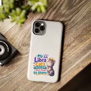 May include: A white smartphone case featuring a graphic of a grey cat wearing a gold crown and a collar with a Libra symbol. The text reads: My Libra Cat's Social skills put mine to shame, accompanied by small colorful stars and hearts.