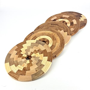 Puede incluir: Cinco posavasos redondos de madera con un diseño tipo mosaico. Los posavasos están hechos de varias tonalidades de madera, incluyendo marrones claros y oscuros, y beige. Cada posavasos tiene un agujero central y un patrón geométrico que irradia hacia afuera.