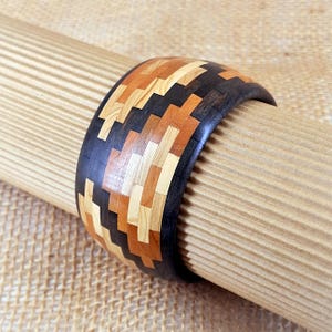 May include: A wooden bracelet with a mosaic design. The bracelet features a pattern of light wood, dark wood, and orange-toned wood pieces. The edges are dark, and the bracelet is displayed on a textured, light brown surface.