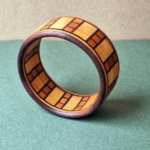May include: A wooden bangle featuring a geometric pattern of brown and light wood squares. The bangle is crafted from dark wood and has a smooth, polished finish.