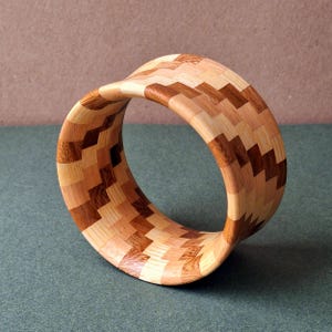 May include: A wooden bangle with a geometric pattern made from different shades of wood. The bangle is curved and has a smooth finish.