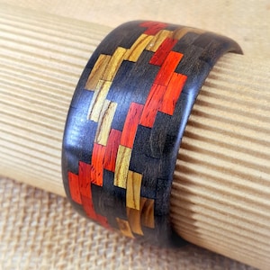 May include: A wide wooden bracelet with a dark finish and inlaid rectangular pieces in red, gold, and orange. The bracelet is displayed on a tan, cylindrical surface, showcasing its curved shape and intricate design. This handcrafted accessory is a unique piece of jewelry.