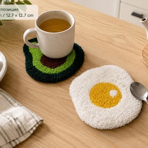 May include: Two tufted coasters measuring 5 x 5 inches. One coaster is shaped like an avocado with green, dark green, and brown yarn. The other coaster is shaped like a fried egg with white yarn and a yellow center. The coasters have a textured surface.