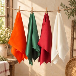 May include: Four square fabric napkins hanging from a horizontal rope with wooden clothespins. The napkins are solid colors: bright orange, deep forest green, dark red, and off-white. Each napkin features a smooth, matte finish and clean edges.