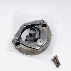 May include: A sewing machine bobbin case with a silver-colored metal hook and a dark gray base. Two small screws are included. The bobbin case is designed to hold the bobbin and thread, essential for sewing.