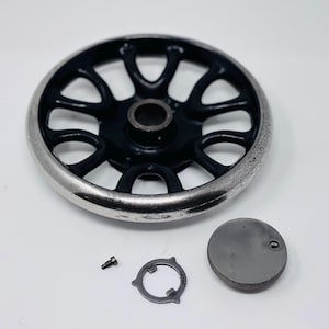 May include: A black and silver sewing machine hand wheel with a silver rim, and a set of small parts. The wheel has a series of spokes and a central hub. Additional parts include a screw, a circular metal piece, and a small, round metal cover.