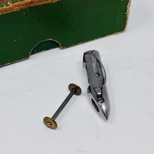 May include: A silver-toned metal sewing machine shuttle with a pointed tip and a curved tension spring, accompanied by a separate metal bobbin featuring a thin central shaft and two small circular brass-coloured end discs for thread storage.