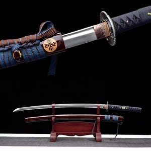 May include: A katana sword with a curved steel blade, a black cord-wrapped handle, and a dark red scabbard featuring a blue cord wrap and a circular emblem. The sword rests horizontally on a dark wooden stand against a black background.