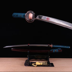May include: A katana sword with a polished steel blade, a teal wrapped handle, and a dark copper-toned circular guard. The sword rests on a black stand above its matching dark red scabbard, which features a teal cord wrap and a gold tassel.