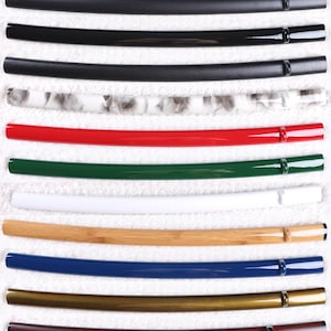 Può includere: Una collezione di undici foderi per spada disposti orizzontalmente. I foderi presentano varie finiture tra cui nero opaco, nero lucido, nero testurizzato, marmo bianco, rosso, verde, bianco, venature del legno naturale, blu, oro metallizzato e legno marrone scuro.
