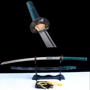 May include: A katana sword with a dark green wrapped handle and a polished steel blade. The black scabbard features white floral detailing and a green cord. A black display stand holds the sword, and a gold tassel rests on a black cloth.