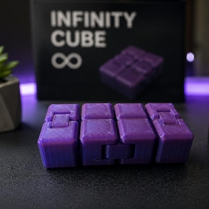 Puede incluir: Un juguete fidget cubo infinito de color morado con un diseño articulado de varios bloques. Al fondo, una caja negra muestra el texto INFINITY CUBE con un símbolo de infinito y una imagen del cubo morado, junto a una pequeña planta suculenta verde.
