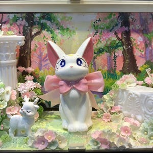 Puede incluir: Una figura de gato blanco con orejas grandes y un lazo rosa, colocada en una vitrina con un pequeño ciervo blanco, una columna blanca, un pájaro blanco y flores rosas y verdes sobre un fondo de bosque pintado.