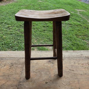 May include: A dark brown wooden stool featuring a contoured, saddle-shaped seat with visible circular joinery details. The stool is supported by four sturdy, cylindrical legs connected by horizontal rungs that provide structural stability.