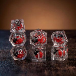 May include: A set of clear polyhedral dice featuring embedded red glitter spheres and small black cat silhouettes. The dice include various shapes with white numbers, including a d20, d12, d10, d8, d6, and d4, arranged in three small stacks.