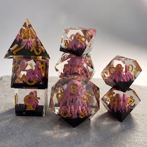 Puede incluir: Un juego de dados poliédricos hechos de resina transparente, cada uno con una pequeña figura de pulpo rosa en su interior. Los dados tienen bordes afilados y números dorados. La base de cada dado está rellena con una capa negra sólida y opaca.