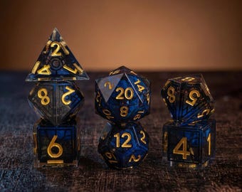 May include: A set of seven polyhedral dice made of translucent dark blue resin with gold-colored numbers. The dice feature a visible internal grid structure and are arranged in three stacks, showcasing various shapes including d4, d6, d8, d10, d12, and d20.