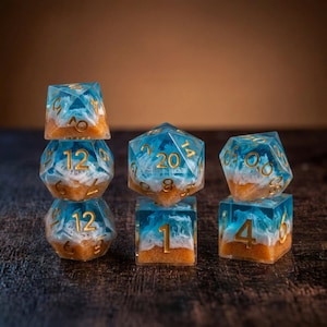 May include: A set of polyhedral dice featuring a translucent blue resin top layer with white swirling patterns resembling ocean waves and a textured sandy-colored base. The numbers are engraved in gold, including a twenty-sided die showing a twenty.
