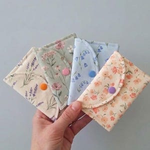 Könnte beinhalten: Vier kleine Stofftaschen mit abgerundeten Klappen, jede mit einem einzigartigen Blumenmuster in Pastelltönen wie Creme, Salbeigrün, Hellblau und Pfirsich. Jede Tasche ist mit einem einzelnen farbigen Kunststoff-Druckknopf in Orange, Pink, Blau oder Violett versehen.
