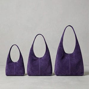 Könnte beinhalten: Drei violette Hobo-Taschen aus Wildleder, die in einer Reihe von klein nach groß angeordnet sind. Jede Tasche verfügt über einen einzelnen integrierten Schulterriemen, eine geschwungene Öffnung und eine vertikale Naht in der Mitte.