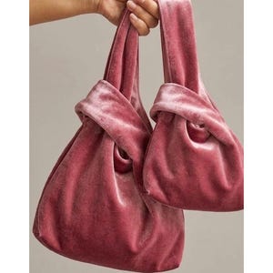 Pode incluir: Duas bolsas de mão de veludo rosa empoeirado com um design de alça com nó. As bolsas são feitas de um tecido macio e felpudo com um brilho sutil. Cada bolsa tem uma alça integrada única que se entrelaça para criar um fechamento seguro.