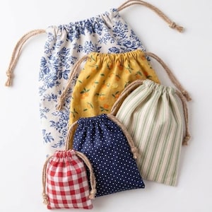 May include: A collection of five fabric drawstring pouches in varying sizes. The bags feature diverse patterns including blue and white floral, yellow with orange fruit, green and white stripes, navy blue with white polka dots, and red and white gingham.