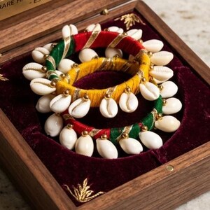 Può includere: Due braccialetti circolari avvolti in filo rosso, verde e giallo con accenti in oro metallizzato, decorati con conchiglie di ciprea bianche. I braccialetti sono esposti all'interno di una scatola di legno con rivestimento in velluto rosso scuro e una targhetta con la scritta RARE HANDCRAFTED JEWELRY - EST. 2024.