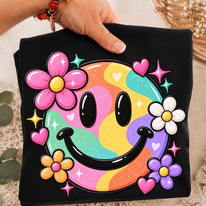 Op de afbeelding: Een zwart t-shirt met een grote, kleurrijke smiley-afbeelding met een golvend, meerkleurig patroon in tinten roze, oranje, geel, groen en blauw. Het ontwerp is omringd door verschillende kleine bloemen en hartjes met fonkelende steraccenten.