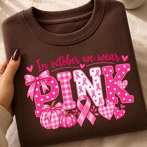Op de afbeelding: Een donkerbruin sweatshirt met ronde hals en een afbeelding met de tekst In october we wear PINK in roze letters. Het ontwerp bevat roze stippen- en vichy-ruitpatronen, een grote strik, twee pompoenen, een bewustzijnslintje en hartjes.