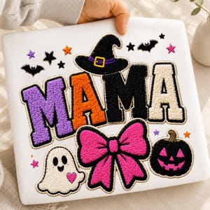 Può includere: Una felpa bianca con la scritta MAMA in grandi lettere testurizzate in stile varsity nei colori viola, arancione e nero. Il design include un cappello da strega nero, un grande fiocco rosa acceso, un fantasma bianco con un cuore rosa e una zucca nera.