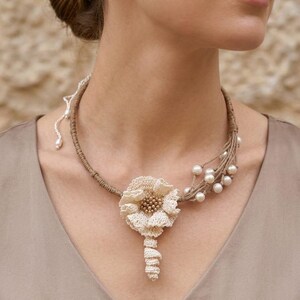 Pode incluir: Um colar de crochê bege com um motivo floral central com contas no centro e uma haste pendente. A banda do colar é feita de cordão enrolado, acentuada de um lado por um grupo de pérolas brancas presas a vários fios finos.