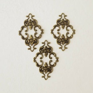 May include: Three antique gold-toned metal decorative embellishments with a floral design. Each embellishment is shaped like a diamond with intricate scrollwork and floral details.