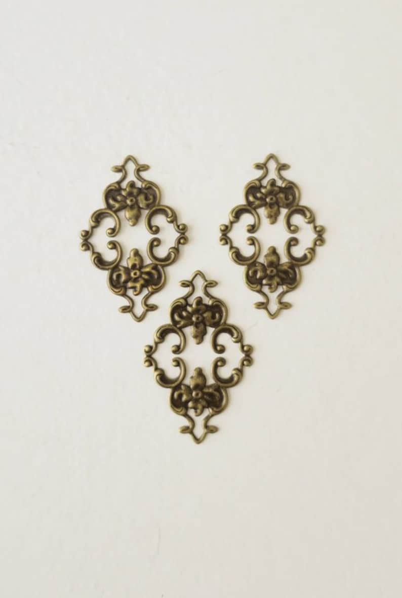 May include: Three antique gold-toned metal decorative embellishments with a floral design. Each embellishment is shaped like a diamond with intricate scrollwork and floral details.