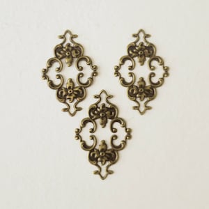 May include: Three antique gold-toned metal decorative embellishments with a floral design. Each embellishment is shaped like a diamond with intricate scrollwork and floral details.