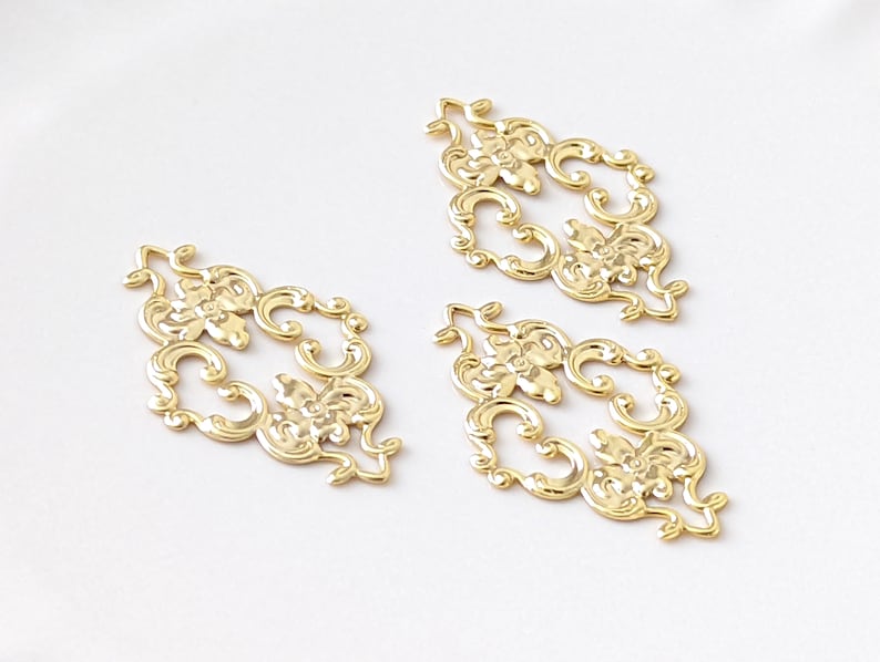 May include: Three gold-colored metal decorative embellishments with a floral design. Each embellishment is shaped like a diamond and has intricate details.