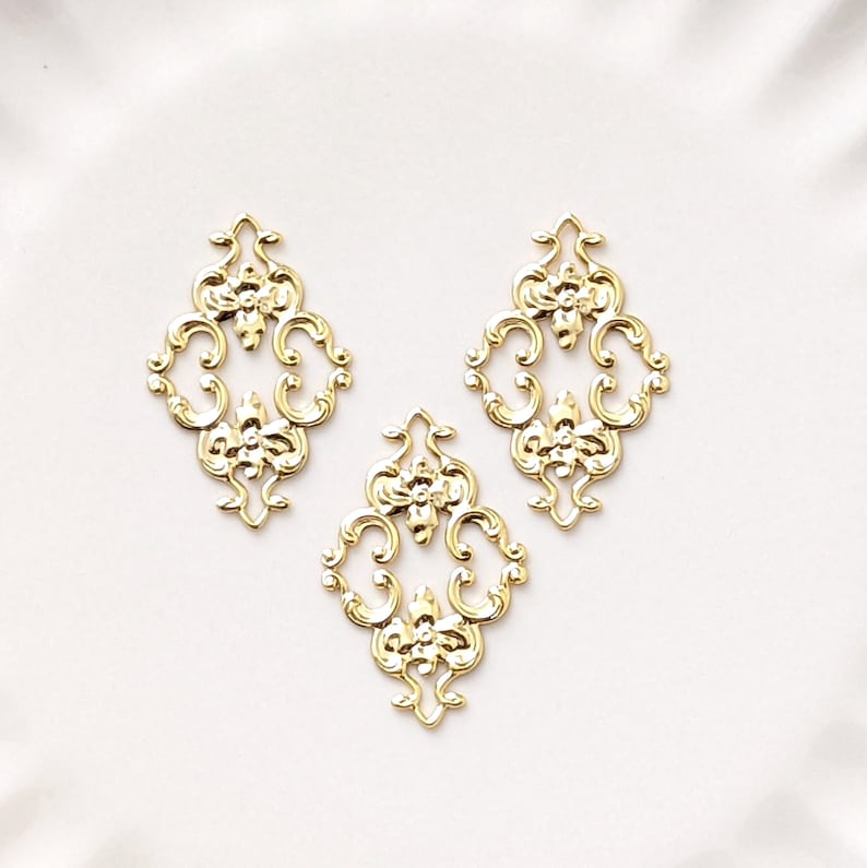 May include: Three gold-colored metal decorative embellishments with a floral design. Each embellishment is shaped like a diamond with intricate scrollwork and floral details.