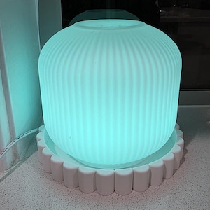 May include: A cylindrical, ribbed, translucent diffuser emitting a bright teal light, resting on a white circular base with a scalloped, rounded edge. A thin white power cord extends from the side of the device, which is illuminated from within.