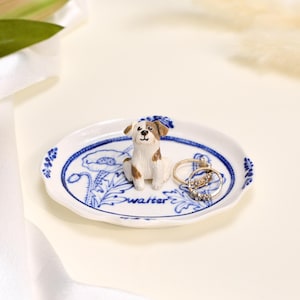 May include: A white ceramic trinket dish with a scalloped edge, featuring blue floral illustrations and the name Walter written in script. A small, three-dimensional brown and white dog figurine sits in the center, accompanied by two gold rings.