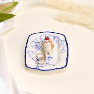 May include: A small white ceramic trinket dish with a scalloped blue rim and blue floral illustrations. A sculpted figurine of a brown and white dog with a red berry on its head sits in the centre. Two gold-toned rings rest on the dish surface.