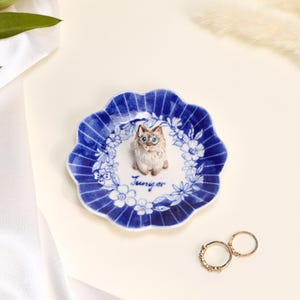 May include: A small, round, scalloped-edge ceramic trinket dish featuring a blue and white floral pattern. A sculpted, hand-painted cat figure sits in the center, with the name "Juniper" written in blue script below it. Two gold-tone rings lie nearby.