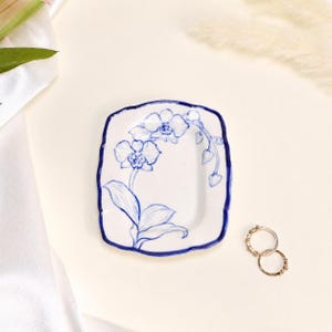 May include: A rectangular ceramic trinket dish with scalloped edges, featuring a hand-painted blue orchid illustration on a white background. The dish has a deep blue rim and is accompanied by two thin gold-toned metal rings placed nearby.